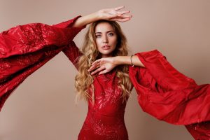 Studio fashion portrait of young blond lovely woman in luxurious red dress with wide sleeves.  Expressive pose. Beige background.